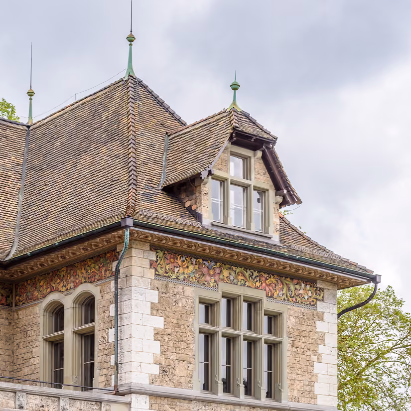 Top section of a historic stone building with steep tiled roof and ornamental window frames.