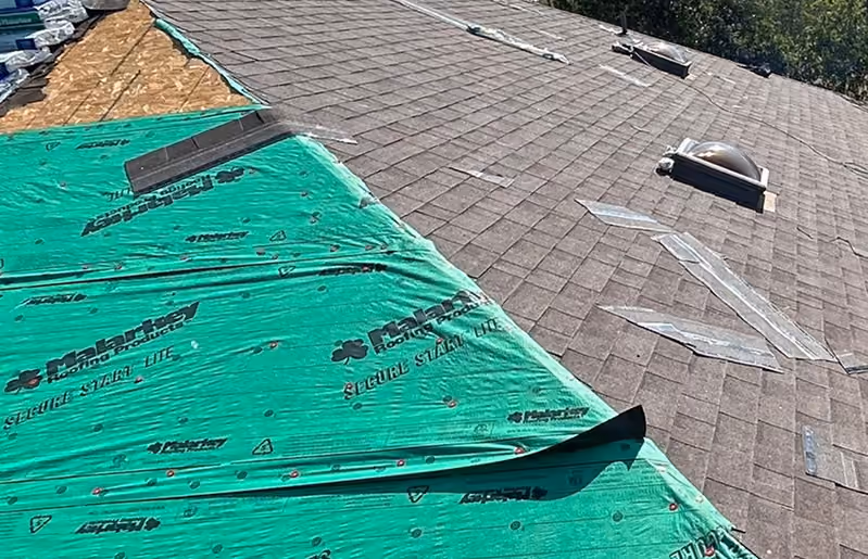 Partially shingled roof with green waterproof underlayment and some exposed plywood sections.