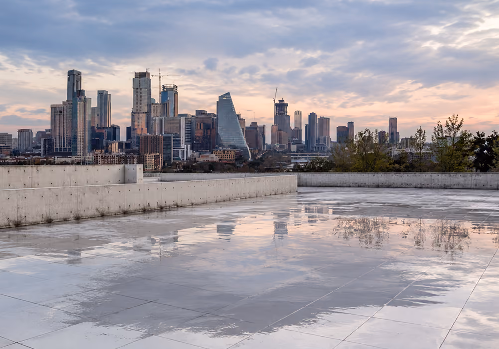Wet rooftop with concrete walls reflecting a city skyline under a cloudy sky at sunset.