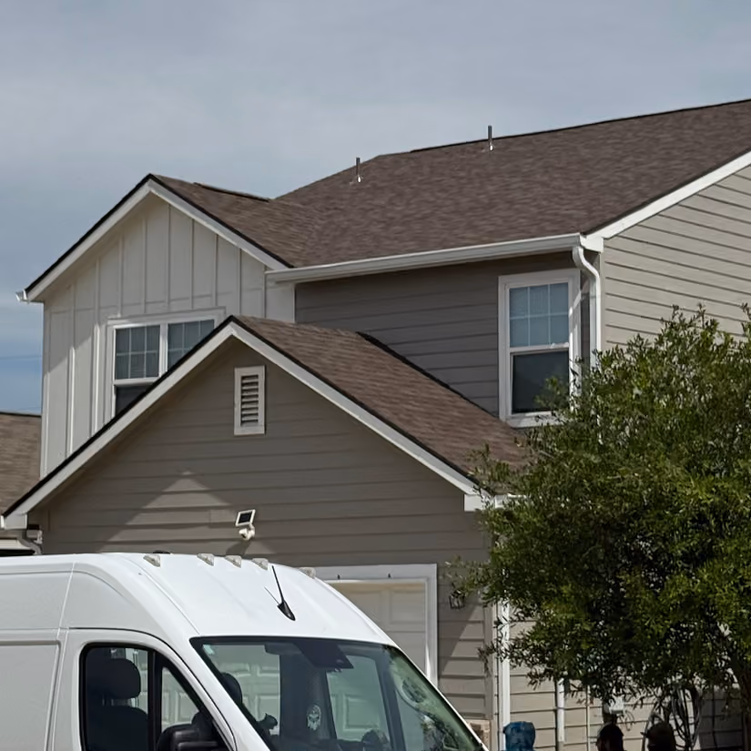 White van parked in front of a beige two-story house with a tree nearby under a cloudy sky.