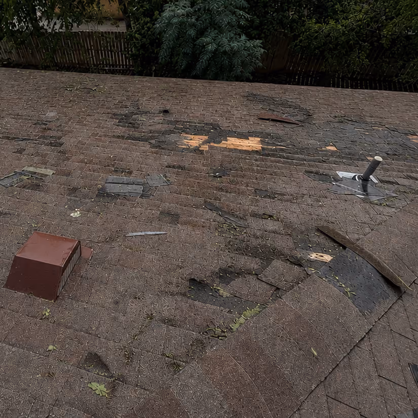 Damaged roof with missing and lifted shingles, exposing plywood underneath surrounded by trees.