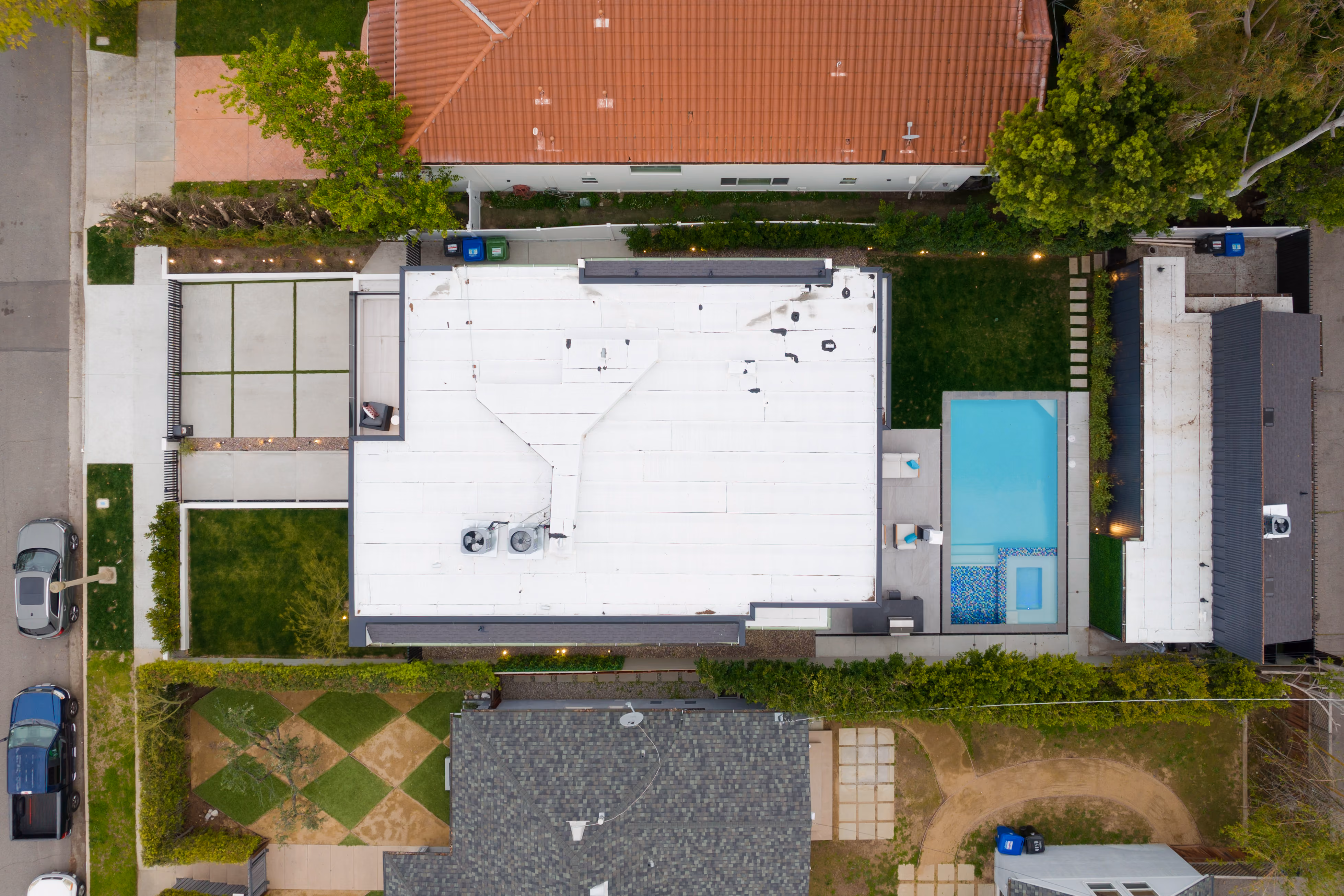 Aerial view of a house with a white roof, swimming pool, driveway, lawn, and neighboring homes.