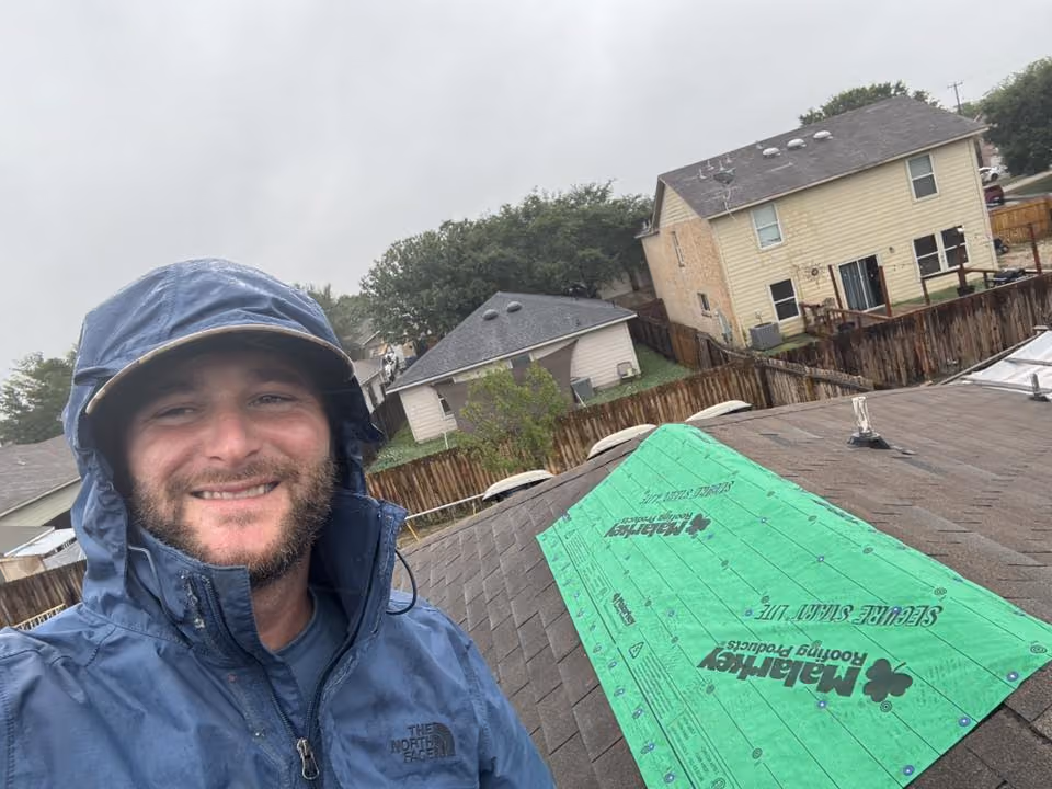 Smiling man in blue rain jacket on roof with green Malarkey roofing underlay on shingles under overcast sky.