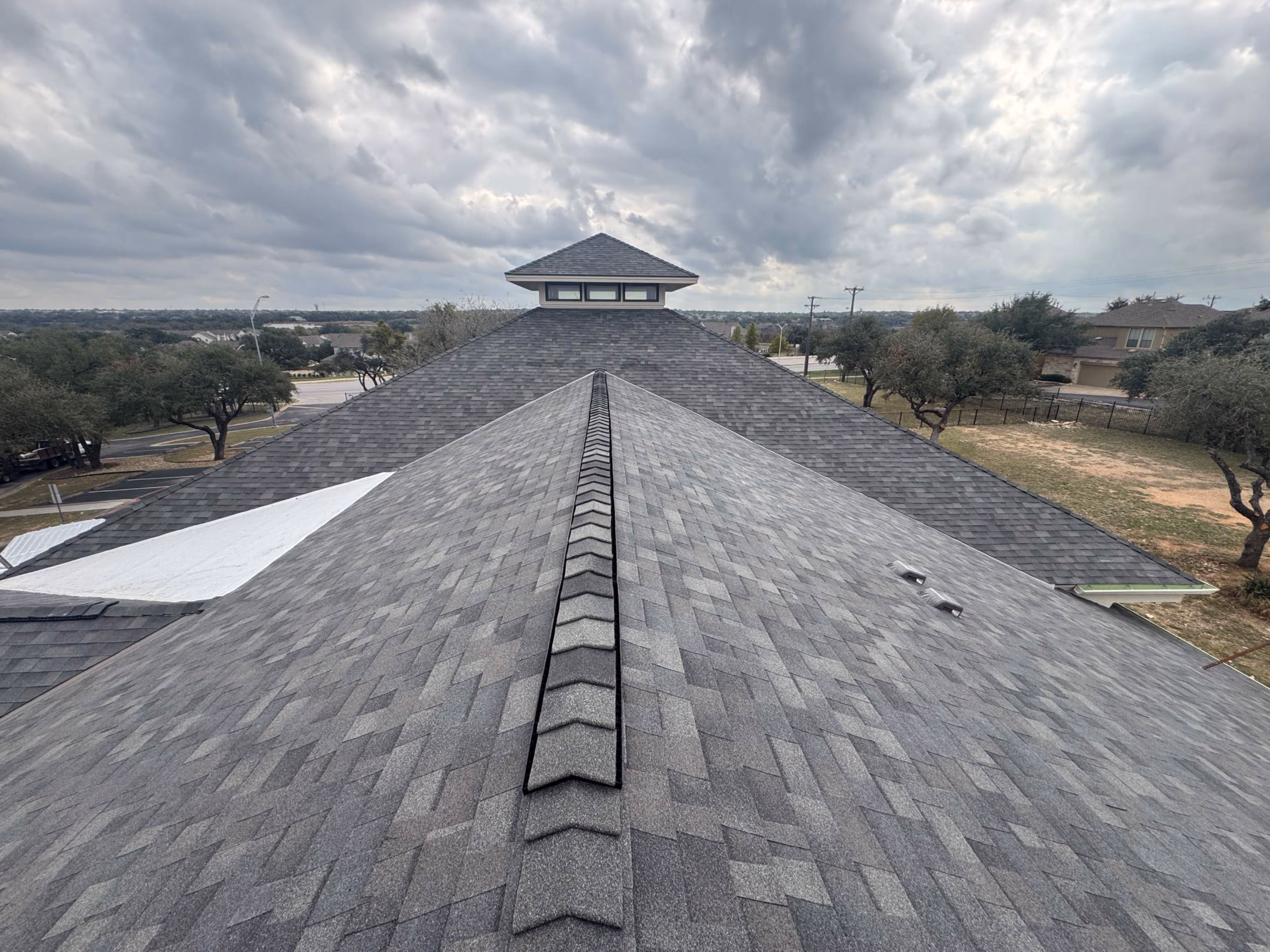 Gray asphalt shingle roof with ridge vent leading to a cupola under a cloudy sky.