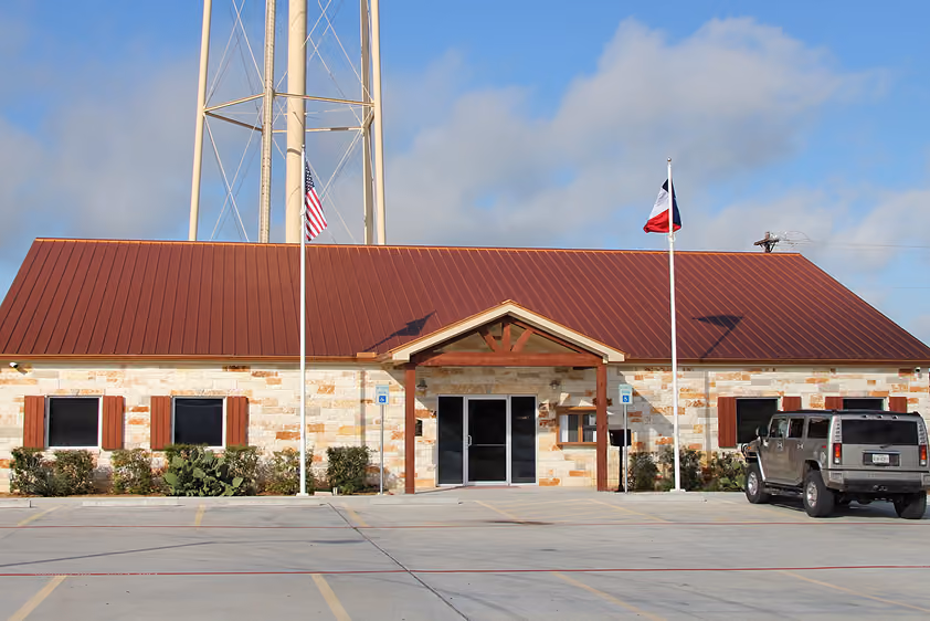 Stone building with red metal roof, two flagpoles with US and Texas flags, and a parked silver SUV.