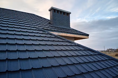 Close-up of black metal roofing tiles with a chimney under a cloudy sky.
