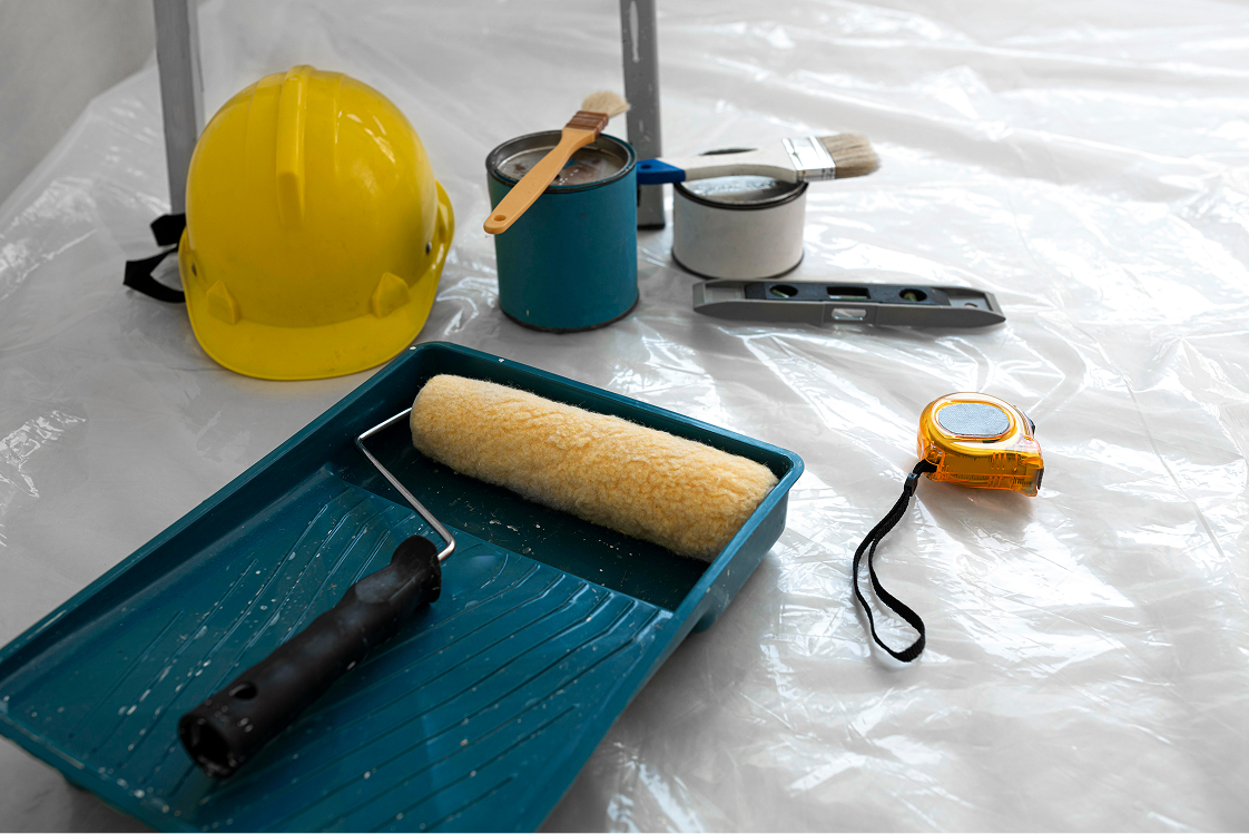 Painting supplies including roller, paint cans, brushes, level, measuring tape, and yellow hard hat on plastic sheet.