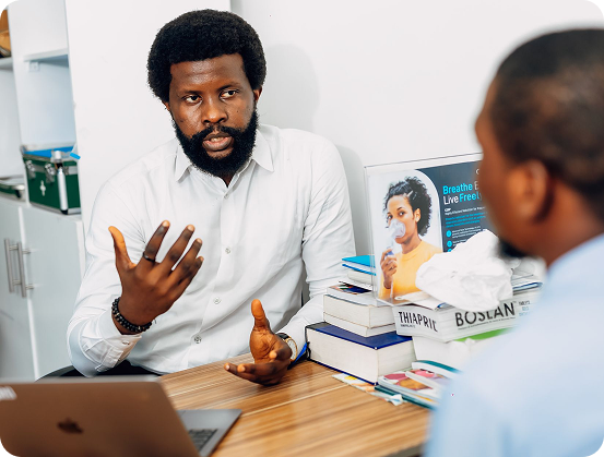 Man in white shirt talking and gesturing at a desk with books and a laptop during a discussion.
