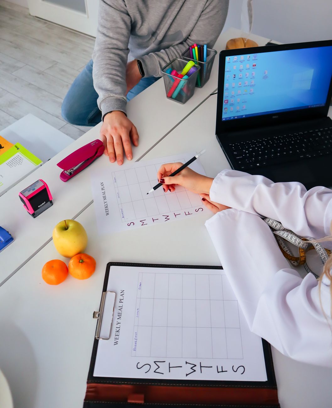 Two people sit at a desk with weekly meal plan sheets, a laptop, fruit, and office supplies.