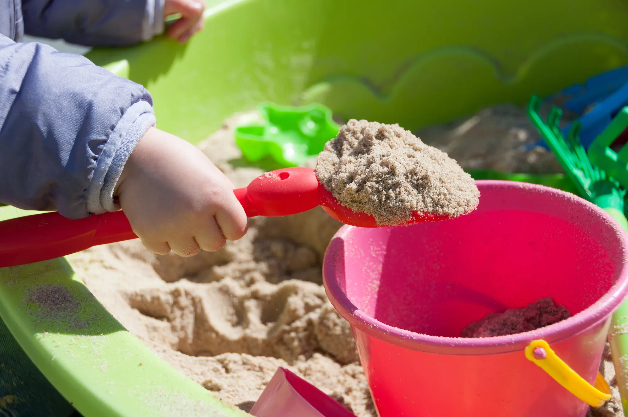 Child scooping sand into a pink bucket with a red shovel