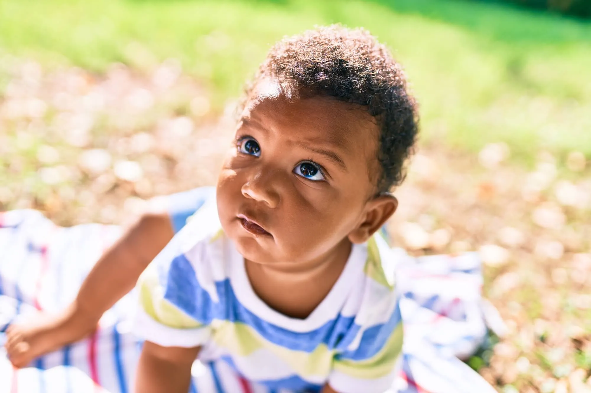 Toddler in a striped shirt looking up while sitting on a blanket outdoors