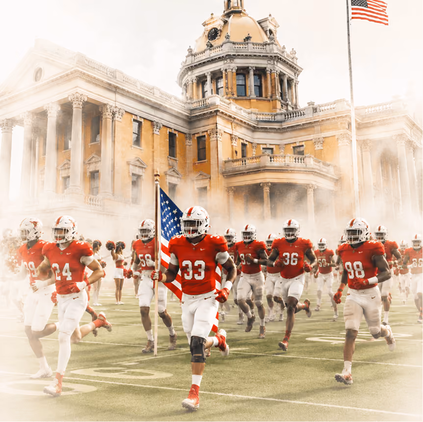 Football players in red and white uniforms run onto the field with an American flag near a large domed building.