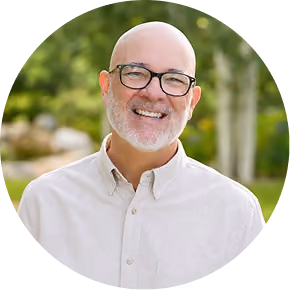 Smiling bald man with glasses and beard wearing light beige shirt outdoors with green trees blurred background.