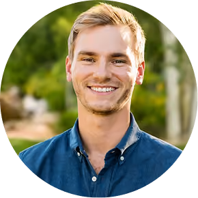 Smiling young man with short blond hair wearing a blue button-up shirt outdoors.