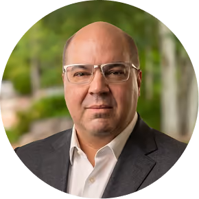 Bald man wearing glasses, white shirt, and dark blazer, posing outdoors with blurred trees behind him.