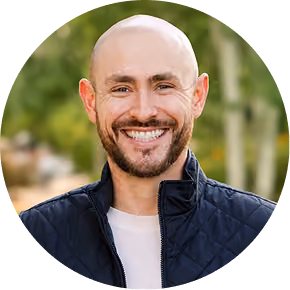 Smiling bald man with beard wearing a black jacket outdoors with blurred green background.
