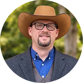 Man with glasses wearing a brown cowboy hat, blue shirt, gray vest, and dark jacket outdoors.