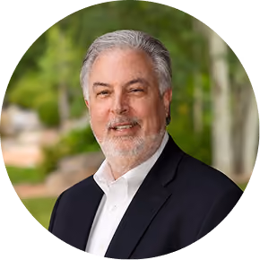 Middle-aged man with gray hair and beard wearing a dark suit jacket and white shirt outdoors.