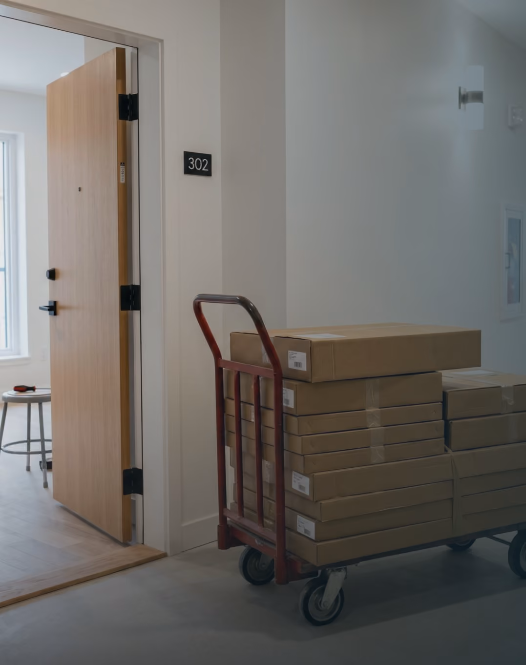 Stack of cardboard boxes on a red hand truck near an open door labeled 302 in a hallway.