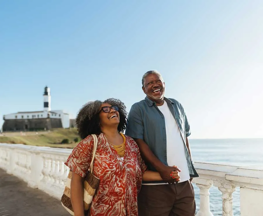 Smiling older couple holding hands by a seaside railing with a lighthouse in the background.