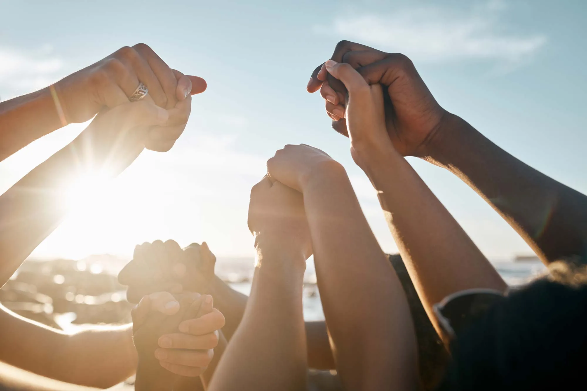 Diverse group of people raising clasped hands together against a bright sky at sunset.