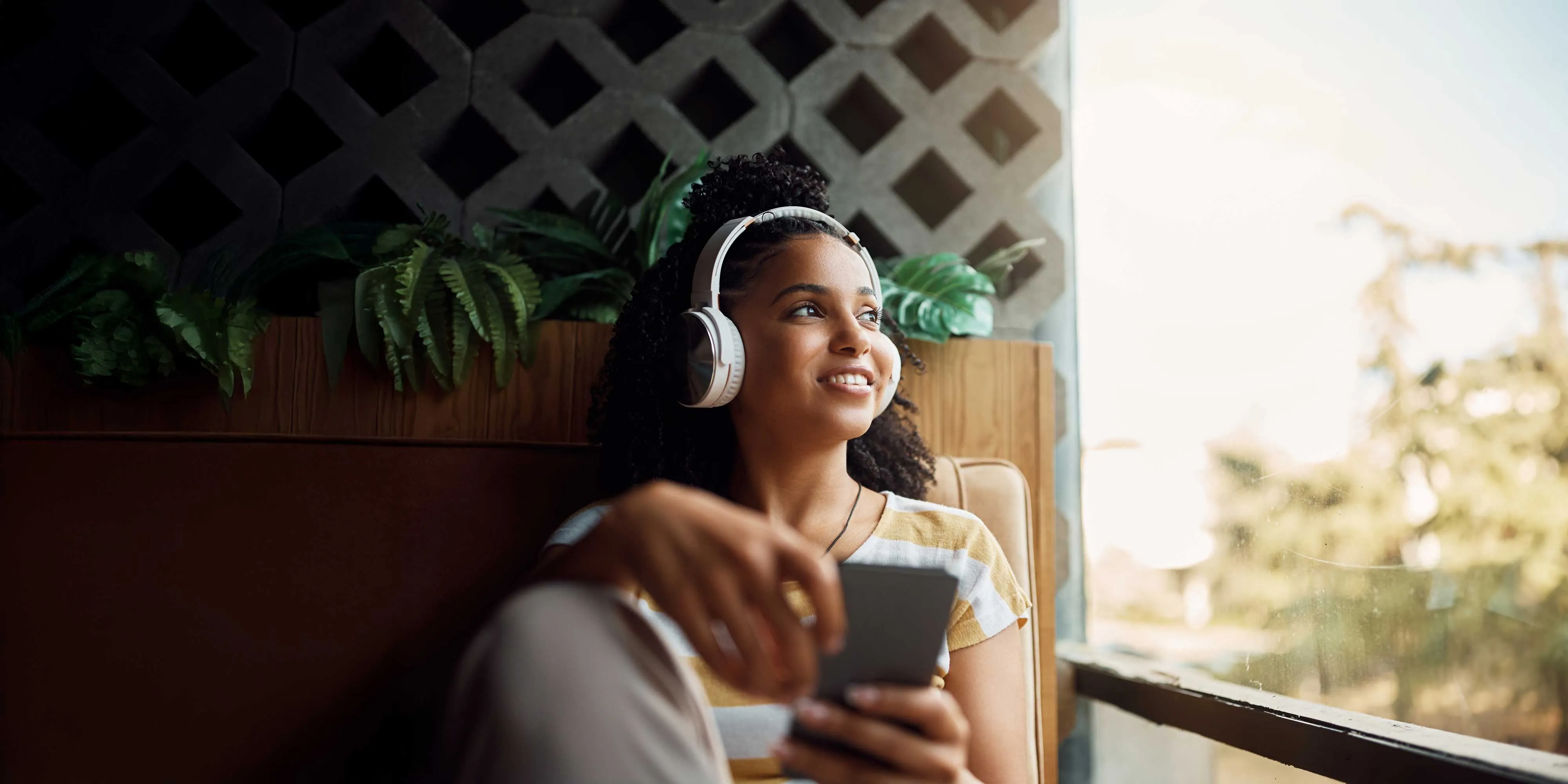 Smiling woman with headphones sitting by a window holding a smartphone, looking outside.