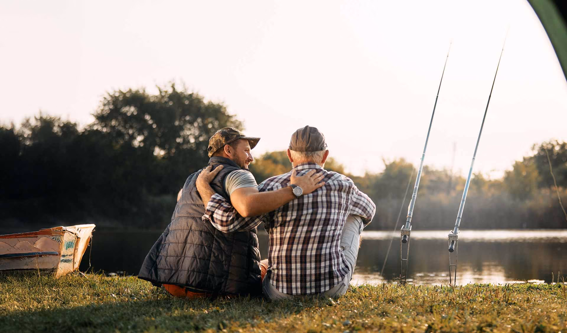 Two men sitting by a lake with fishing rods, one with arm around the other's shoulder.