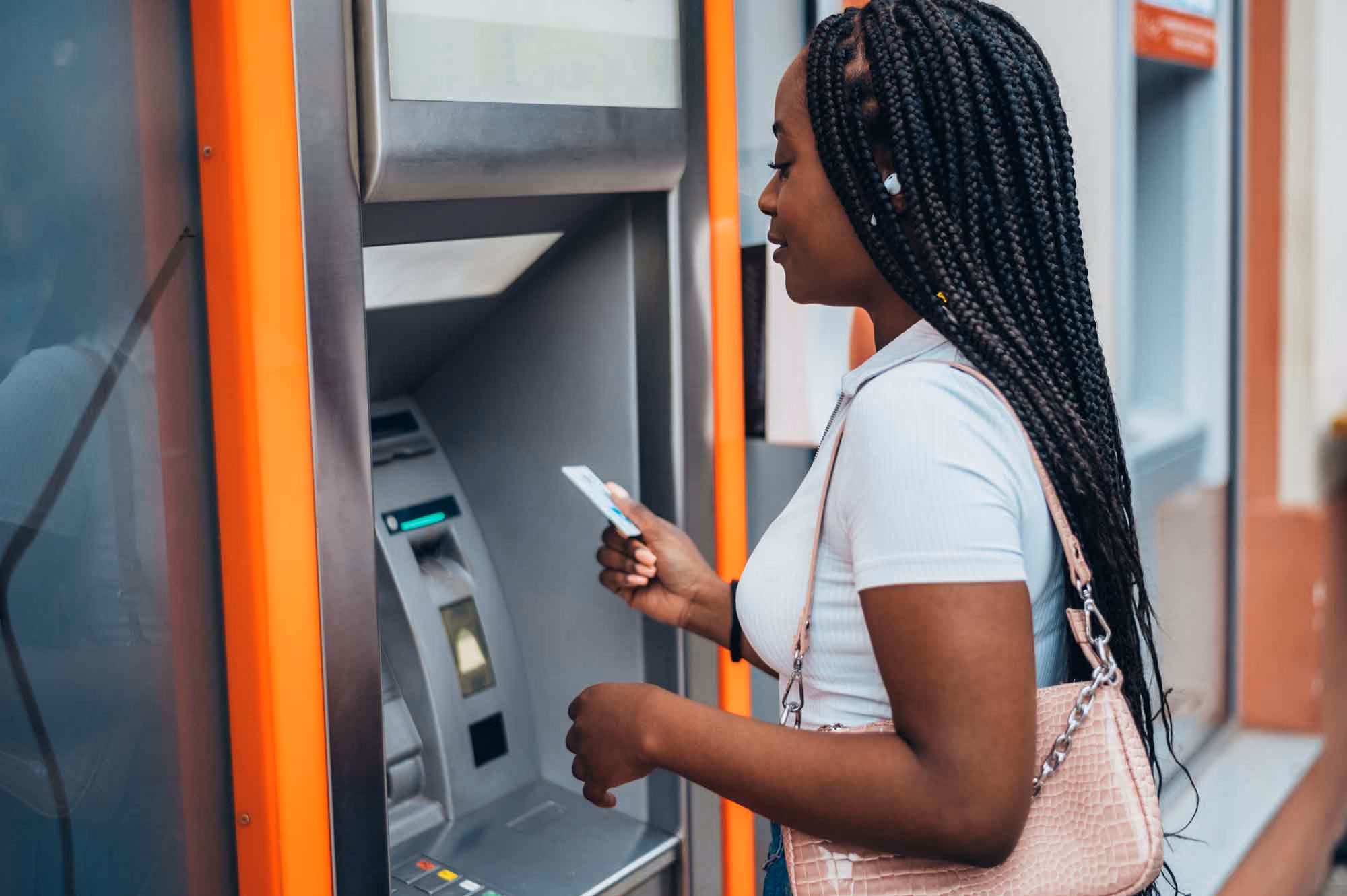 Woman with braids holding a card at an outdoor ATM machine with orange accents.