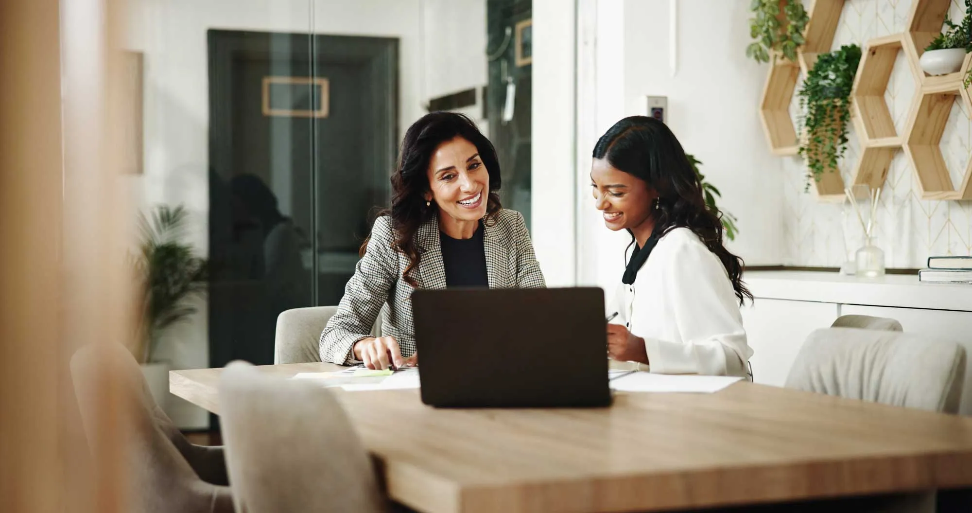 Two women smiling and working together at a table with a laptop and papers in a modern office.