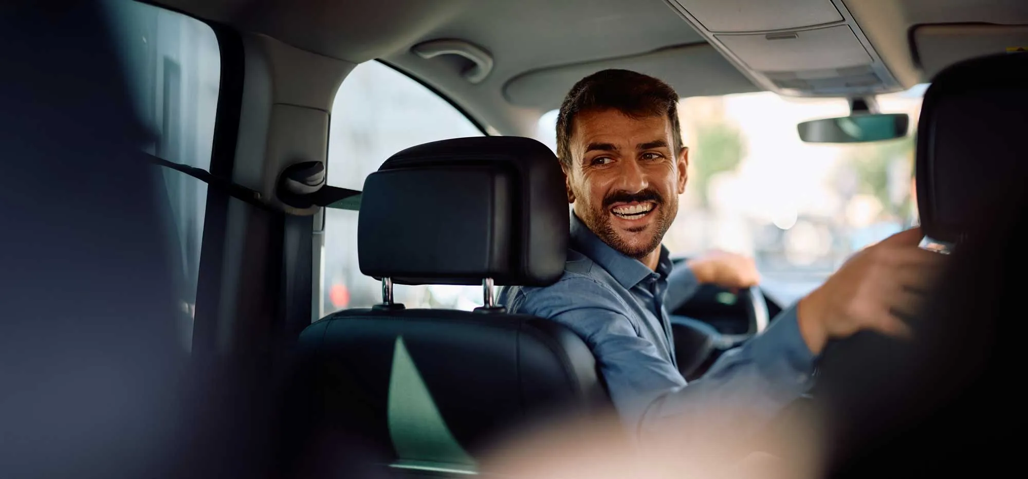Smiling man in blue shirt driving a car and looking back at the passenger seat.