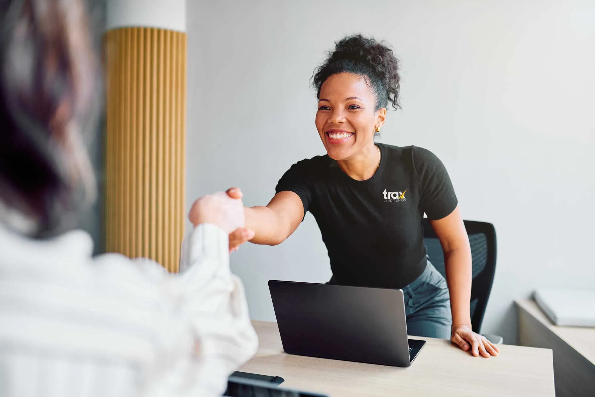 Smiling woman in black shirt with Trax logo shakes hands with a person across a desk with a laptop.