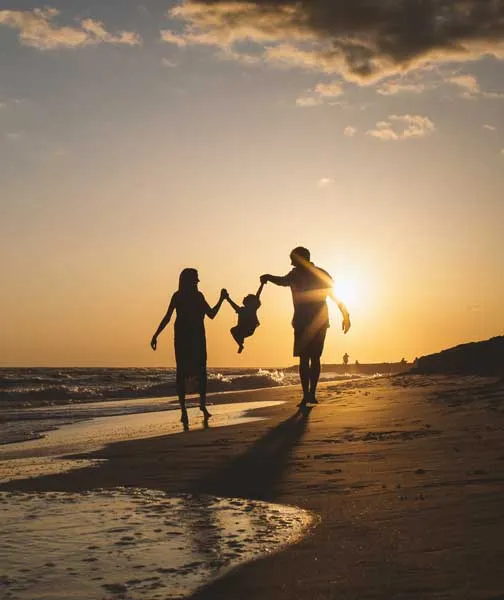 Silhouettes of two adults swinging a child by the arms on a beach at sunset.