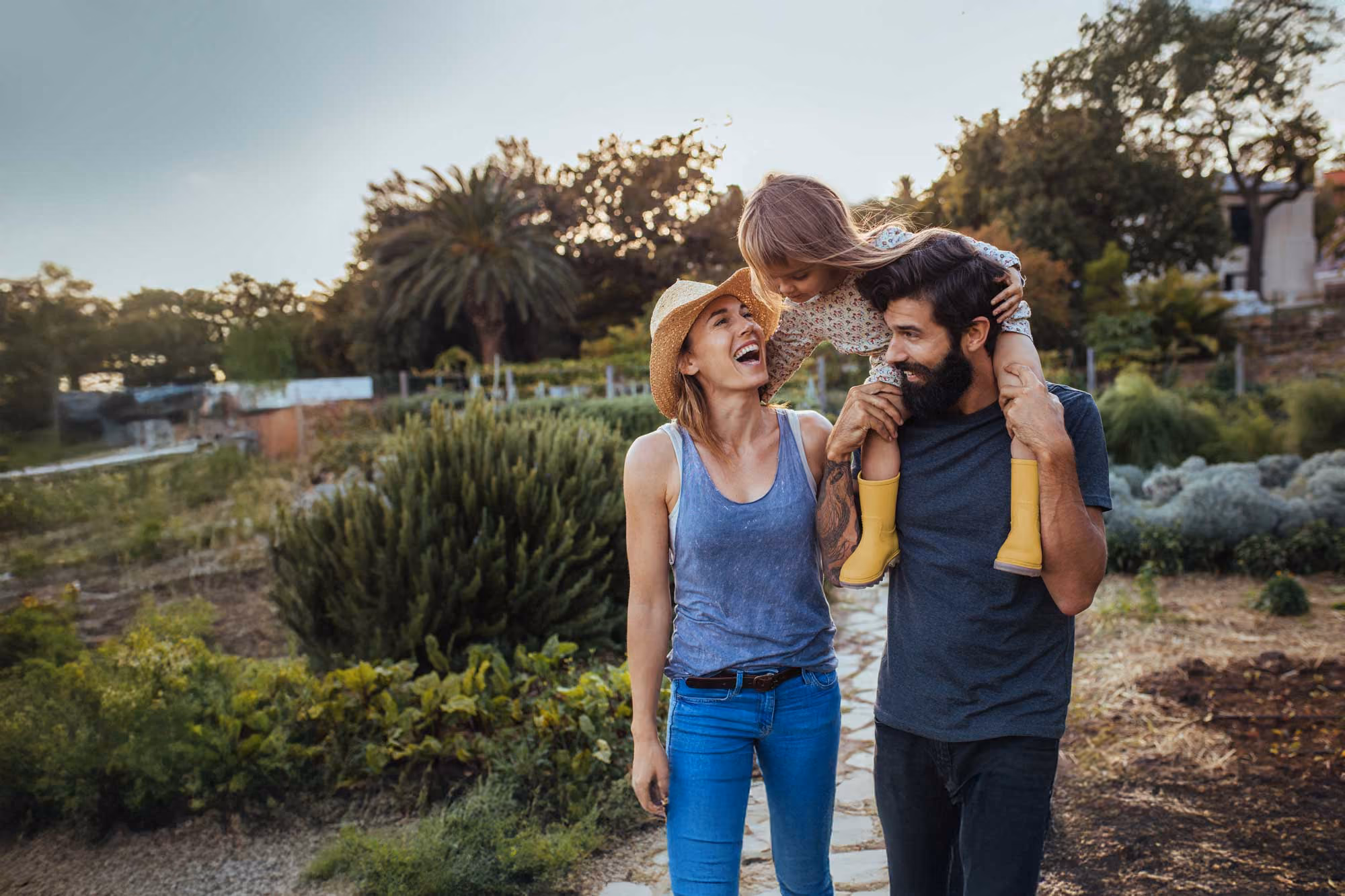 Happy family outdoors: mother smiling, father carrying child with yellow boots on shoulders.