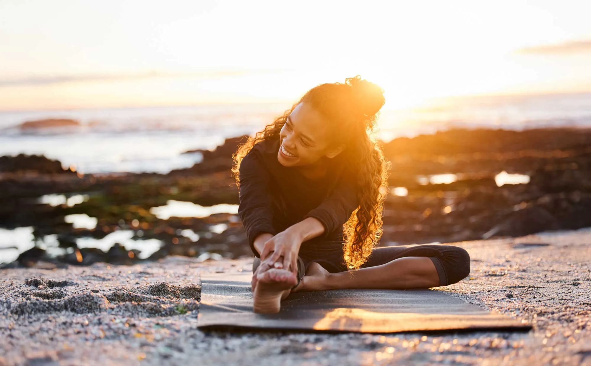 Woman stretching on a yoga mat on a rocky beach at sunset, smiling and reaching for her foot.
