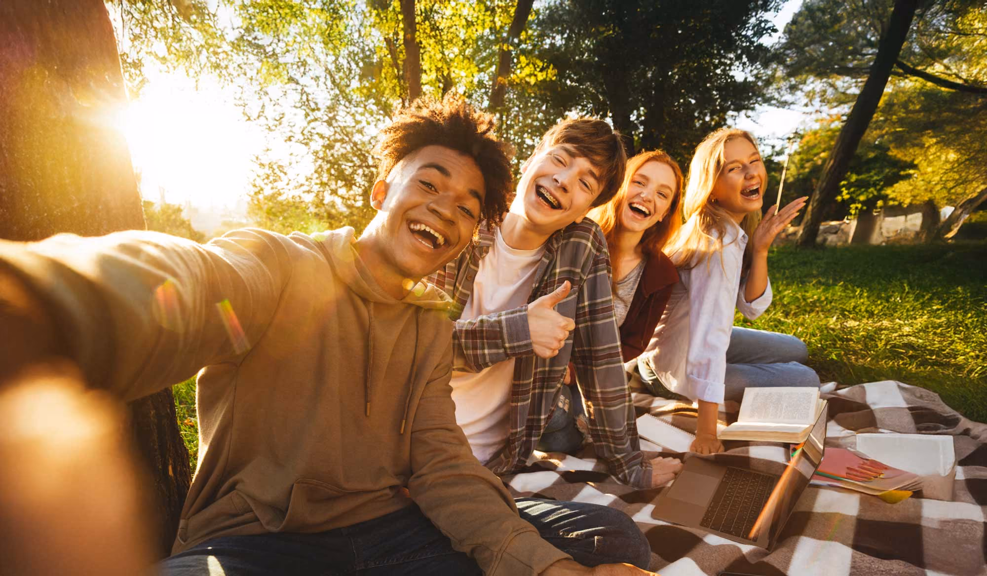 Four teens happily sitting on a blanket outdoors with books and a laptop, taking a selfie.