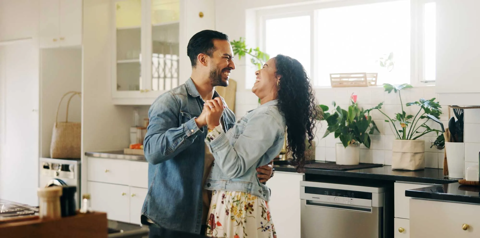 Happy couple holding hands and dancing in a bright, modern kitchen with plants and appliances.