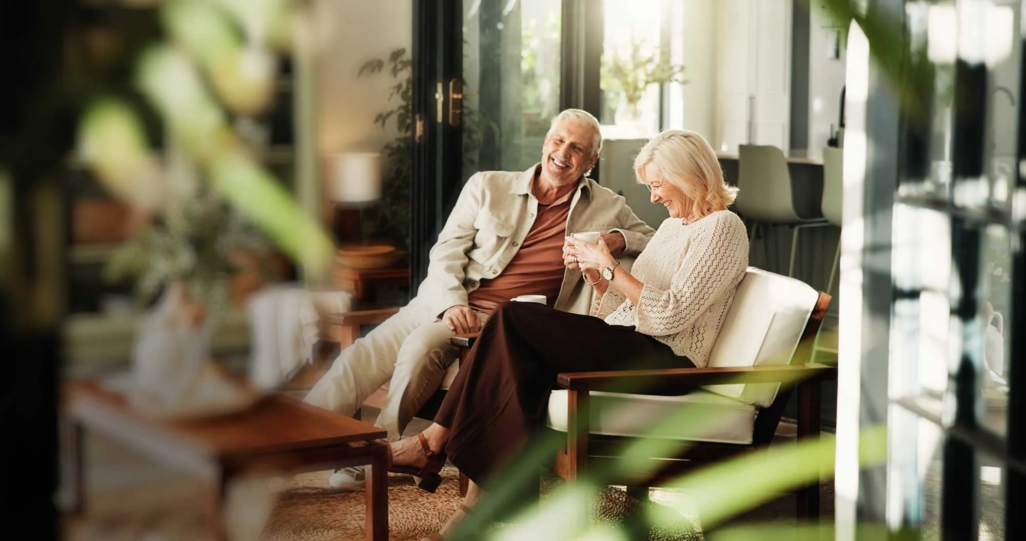 Smiling elderly couple sitting and laughing on a cozy patio with plants around them.
