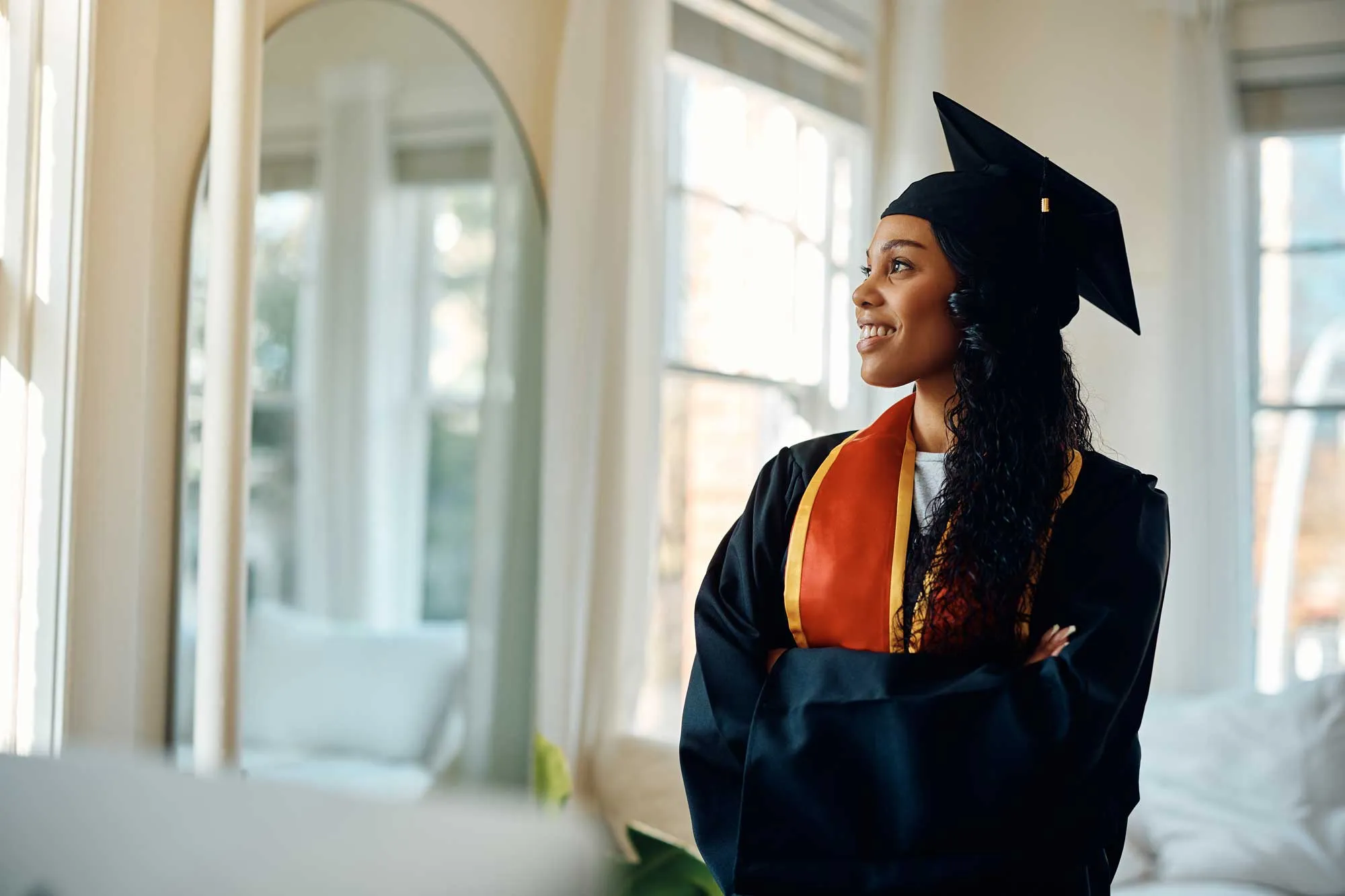 Smiling woman in graduation cap and gown standing indoors with arms crossed, looking out a window.