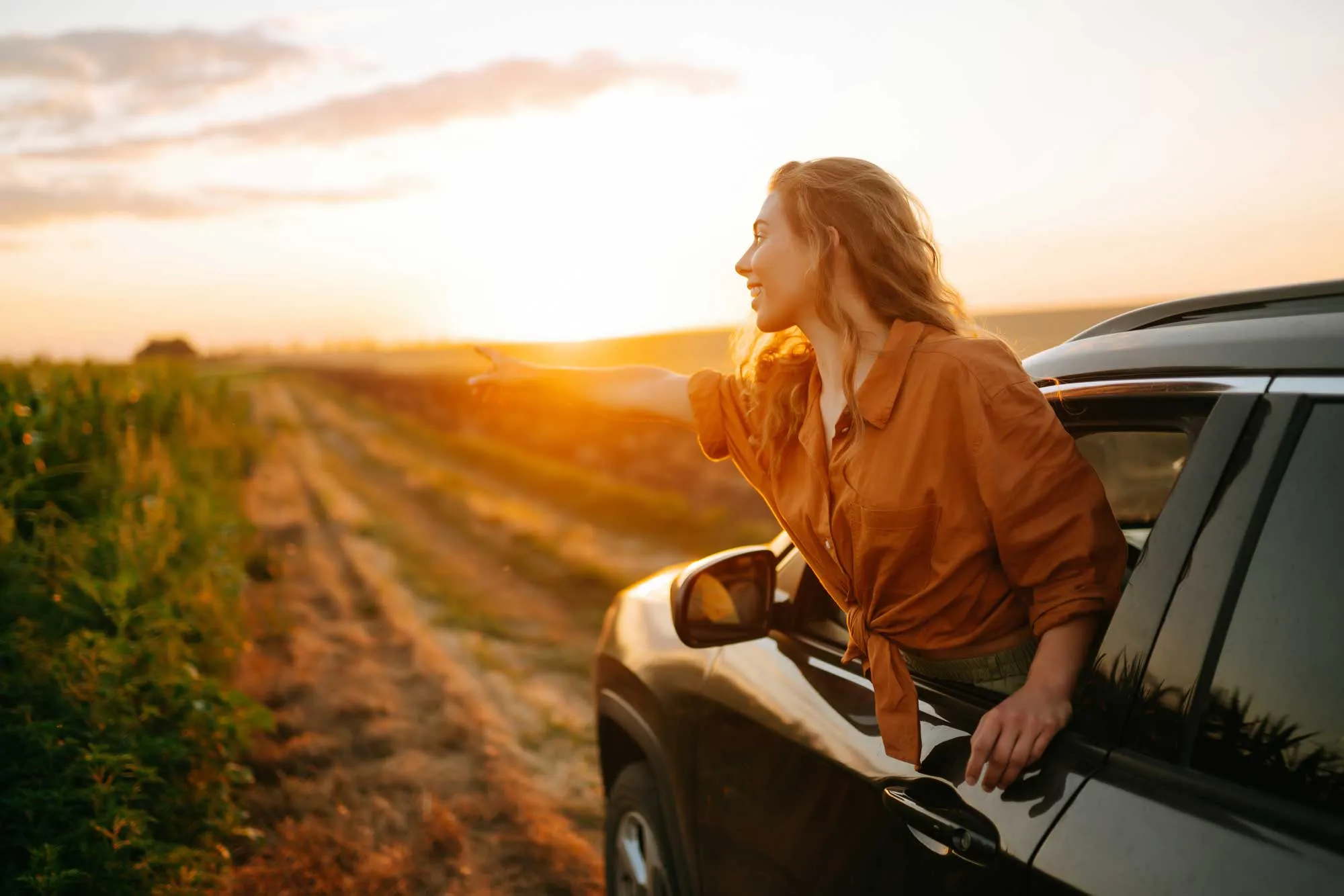 Woman leans out of car window pointing towards sunset over a dirt road and fields.