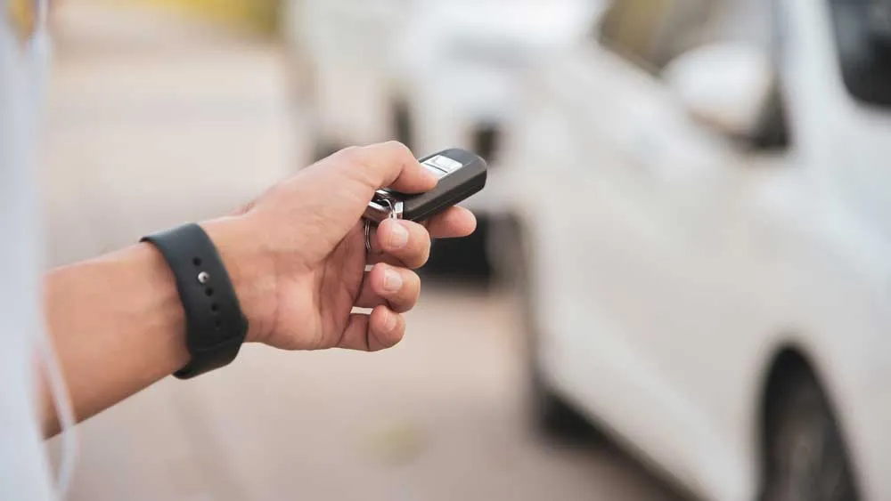 Hand holding a car key fob pointed towards a white car for remote unlocking or locking.