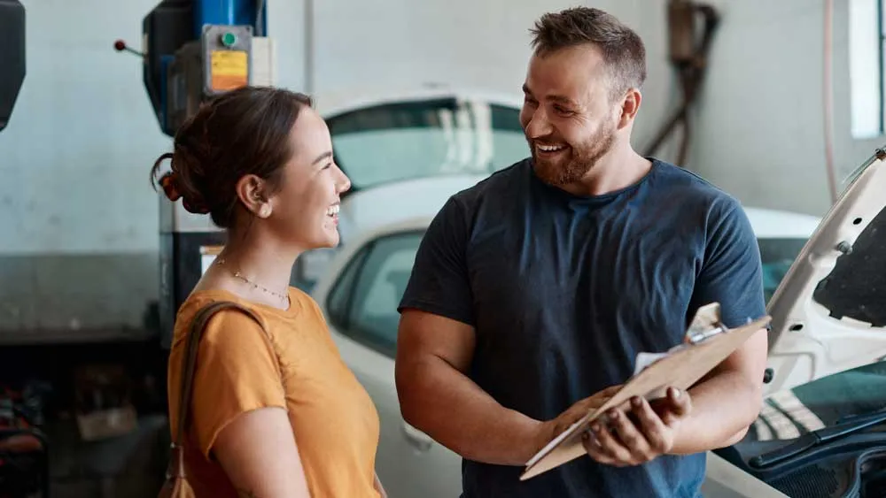 Mechanic smiling and holding clipboard talking to a female customer in a car repair shop.