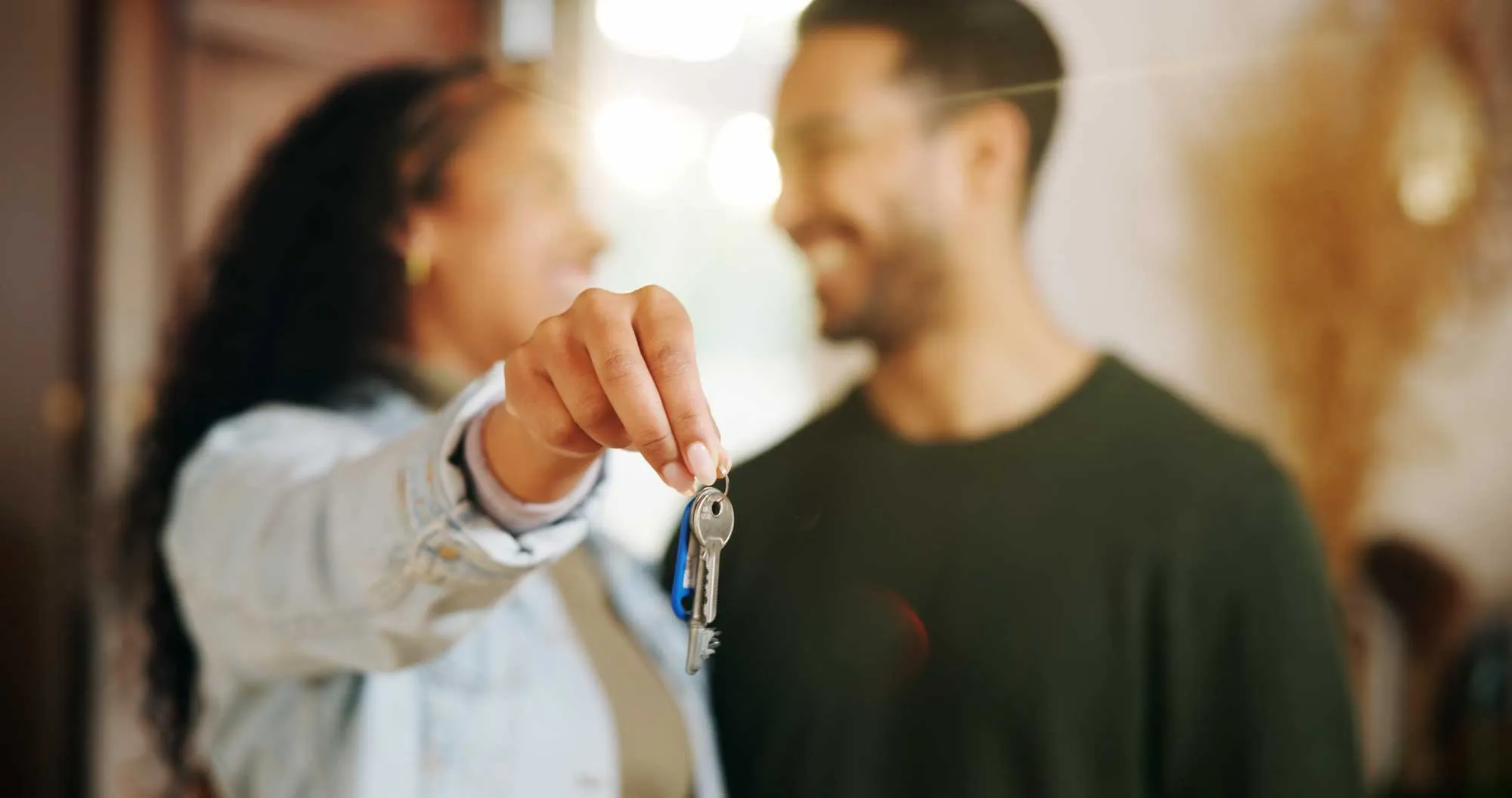 Woman holding house keys toward the camera with man smiling in the background indoors.
