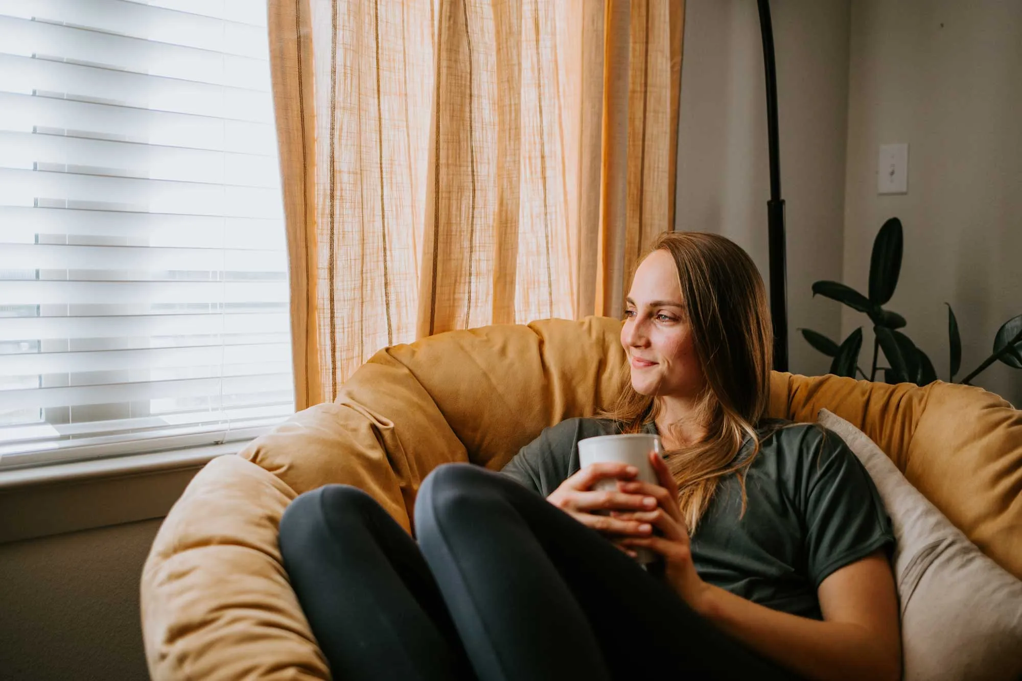 Woman sitting on a cushioned chair near a window holding a white mug, looking outside and smiling.