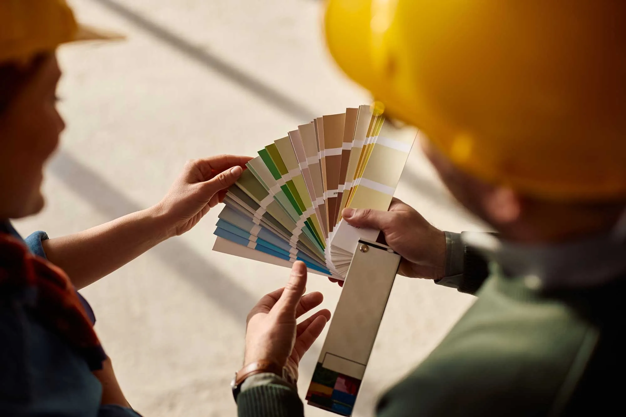 Two people in yellow helmets examining a fan of paint color swatches indoors.