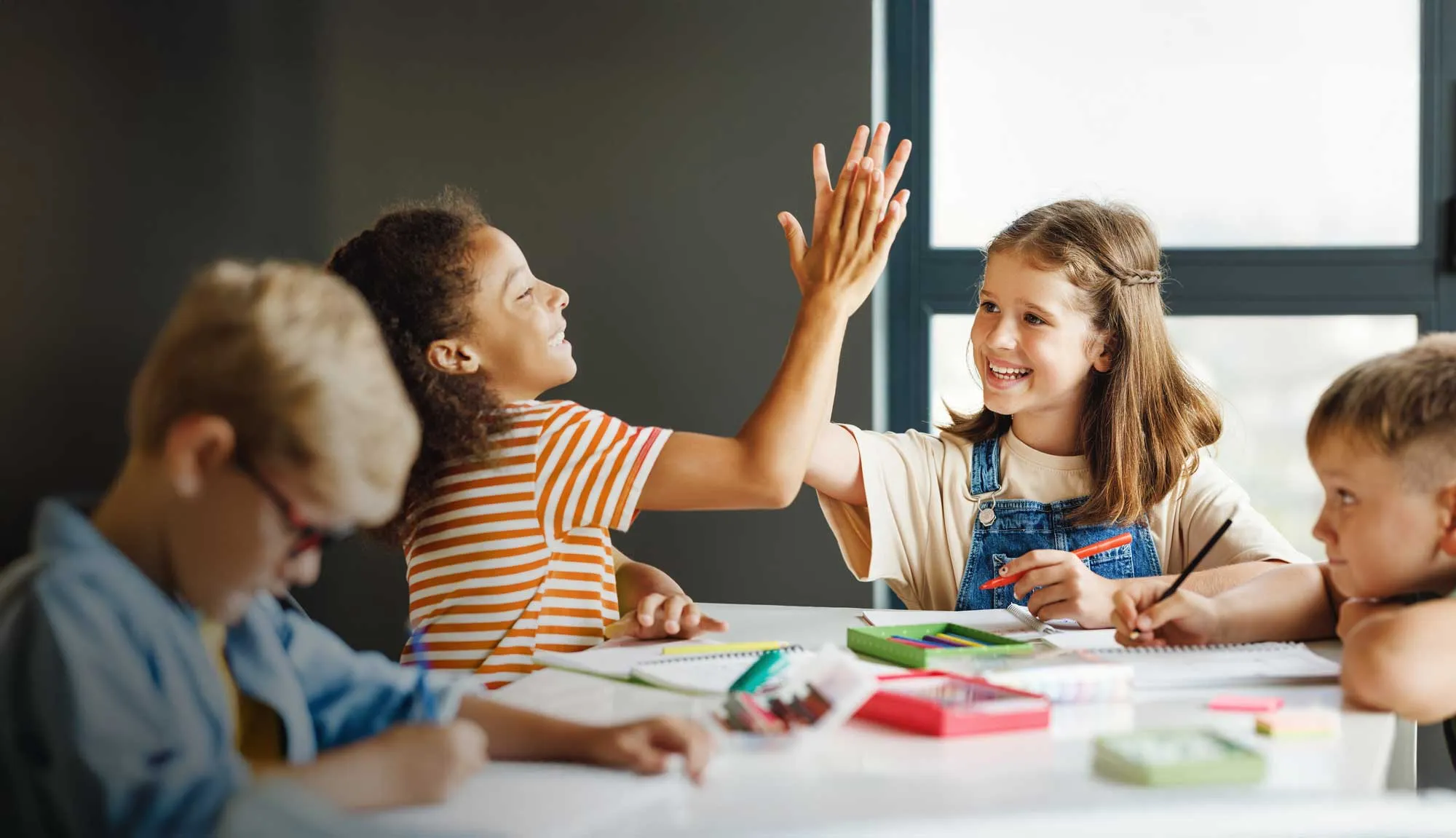 Two girls smiling and giving a high-five while sitting at a table with classmates doing homework.