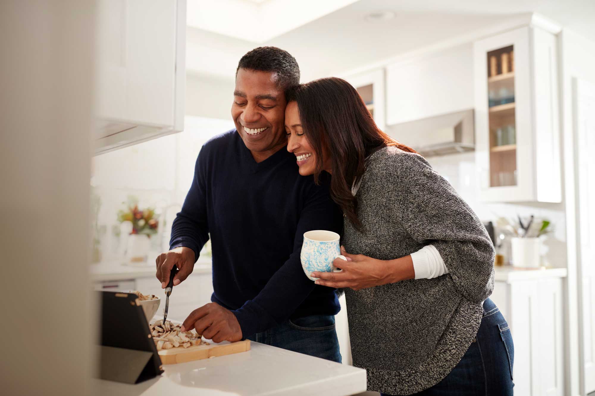Smiling couple preparing food together in a bright kitchen, woman holds a mug while man chops ingredients.