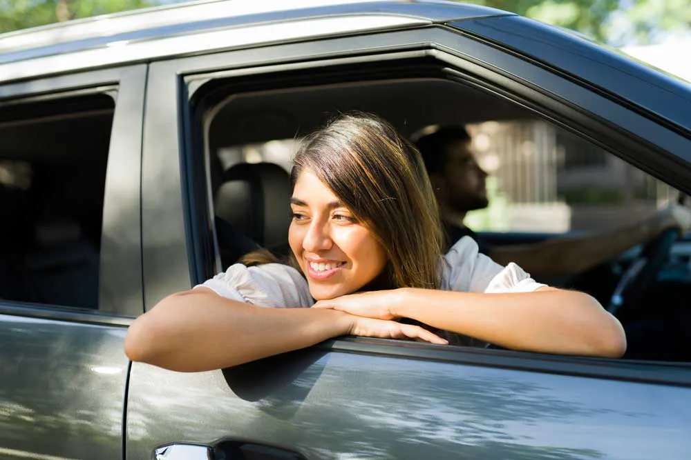 Smiling woman leaning out of a car window with a man driving in the background.