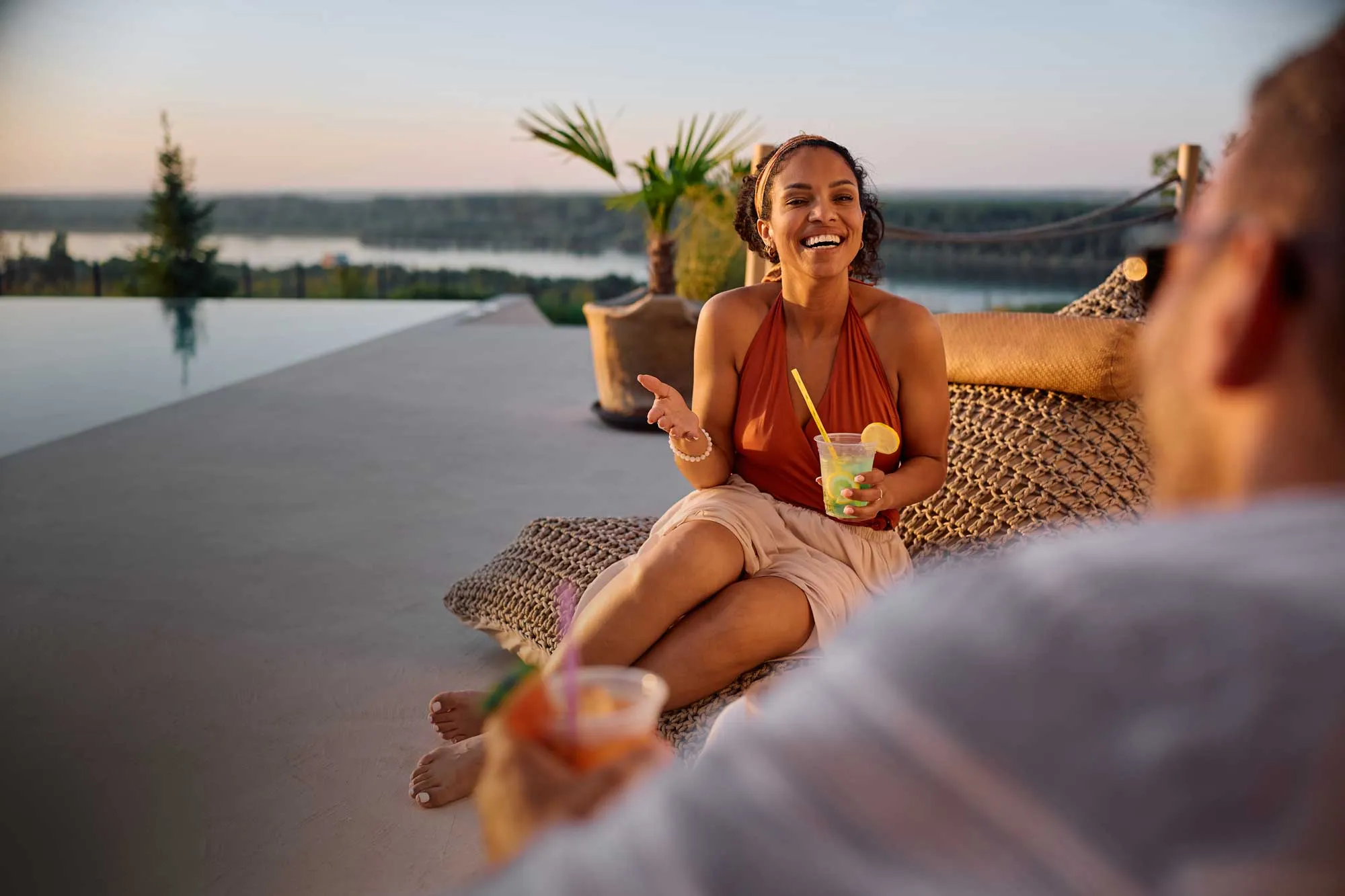 Woman in orange top sitting on wicker chair by pool, smiling and holding lemonade with lemon slice.