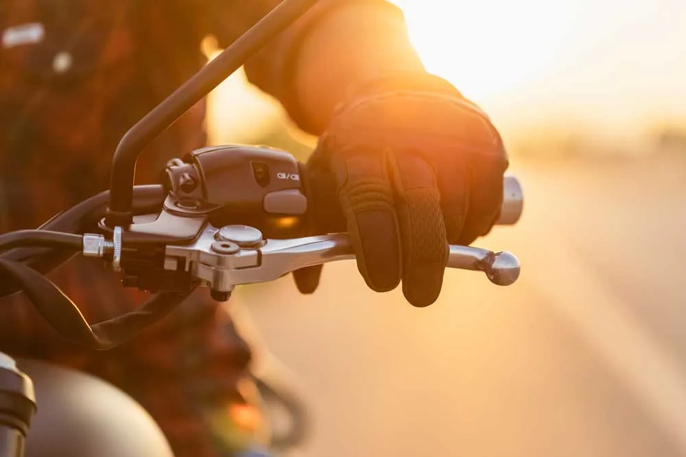 Gloved hand gripping motorcycle handlebar with sunlight in the background.