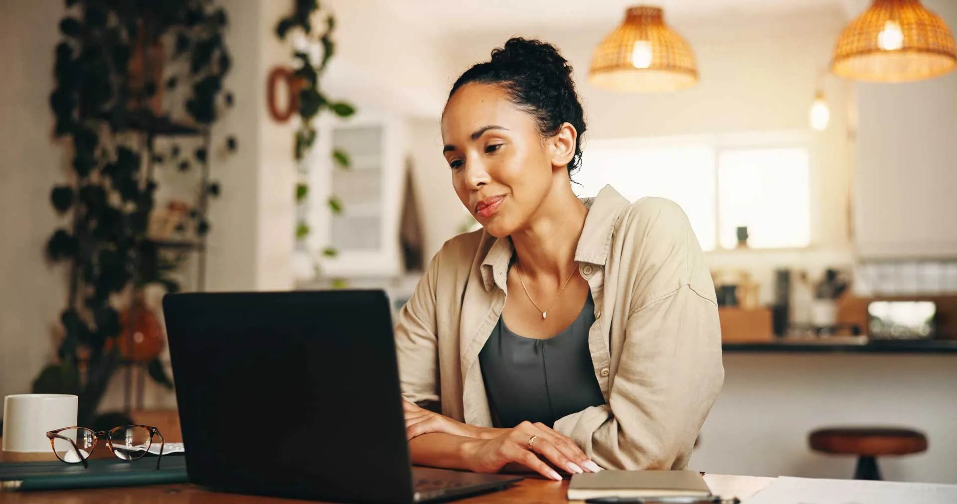 Woman with hair in a bun using a laptop at a wooden table in a warmly lit home setting.